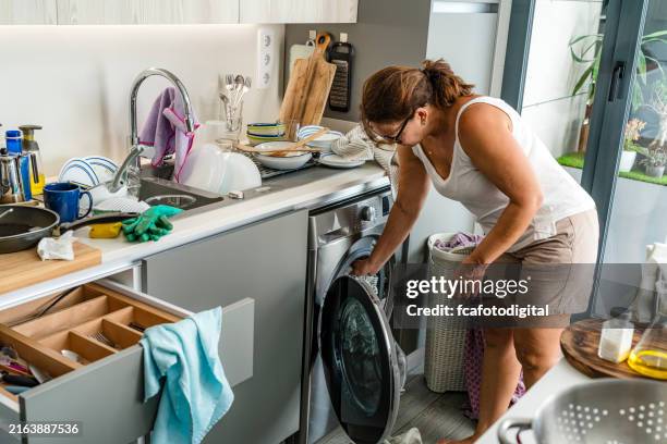 woman loading clothes in washing machine in a messy kitchen - tired housewife stock pictures, royalty-free photos & images