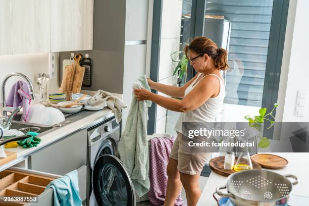 mujer cargando ropa en la lavadora en una cocina desordenada - tarea doméstica fotografías e imágenes de stock