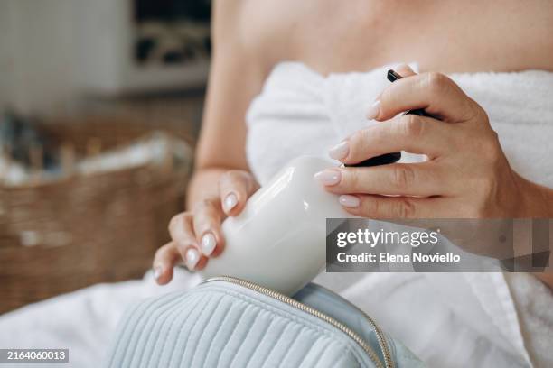 female hands takes a white bottle of moisturizing or anti-aging body lotion from a cosmetic bag, the woman is wrapped in a towel after showering - anti age stock-fotos und bilder