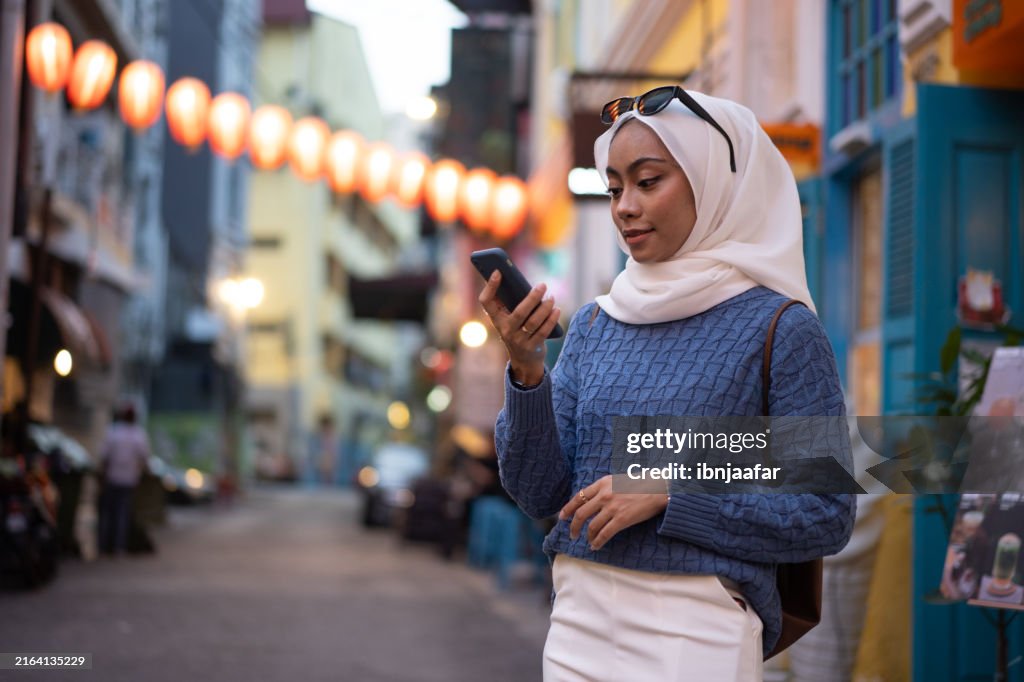 Mujer joven con hiyab usando un teléfono inteligente mientras camina en una calle muy iluminada con linternas colgantes durante la noche