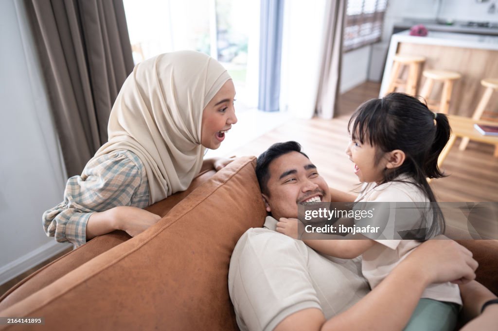 A happy family of three, with a mother wearing a hijab, enjoys a playful moment on the sofa in their modern living room