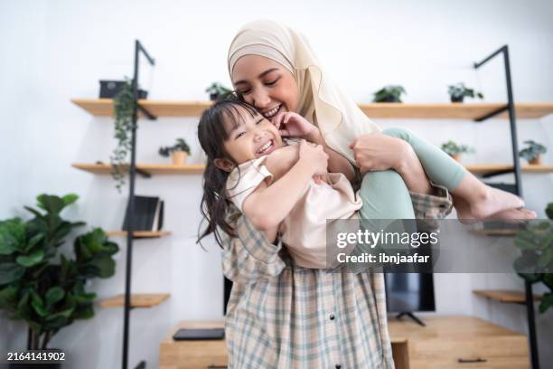 a happy mother wearing a hijab is joyfully hugging her smiling young daughter in a cozy, sunlit living room with plants and a tv in the background - islam stock pictures, royalty-free photos & images