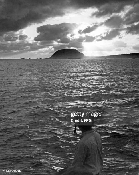 Rear view of an American military officer looking out over the Pacific Ocean, naval vessels in silhouette on the horizon, during the Battle of Iwo...