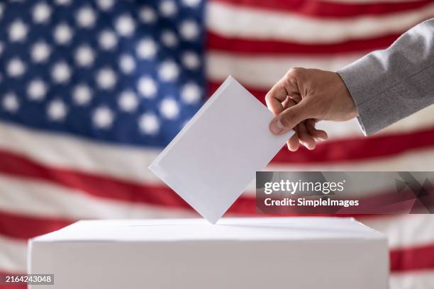 election in usa. man hand putting his vote into ballot box and american flag on background - close up - election stock pictures, royalty-free photos & images