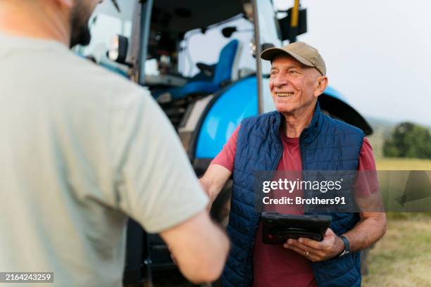 smiling senior farmer with tablet shaking hands with man on farmland - agricultor imagens e fotografias de stock