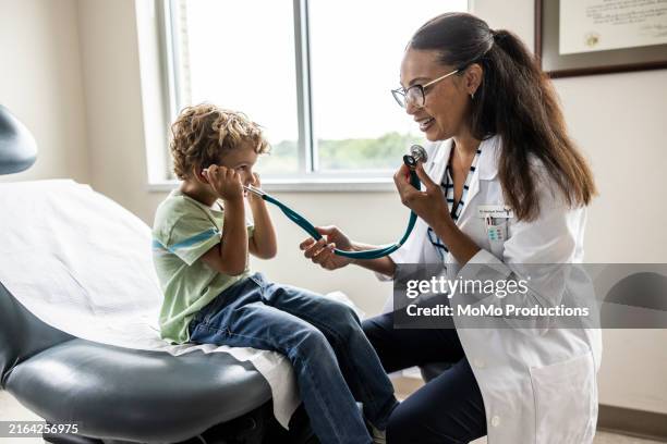 female doctor letting young boy listen to stethoscope in exam room - hospital infantil fotografías e imágenes de stock