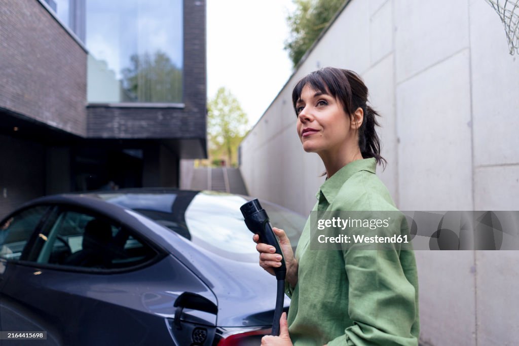 Mature businesswoman holding electric plug and standing near car in parking