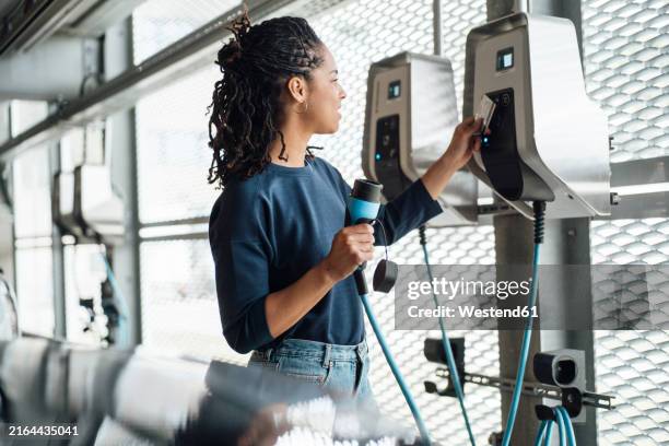 smiling young businesswoman paying through credit card holding electric plug at parking garage - borne de recharge pour véhicule électrique photos et images de collection