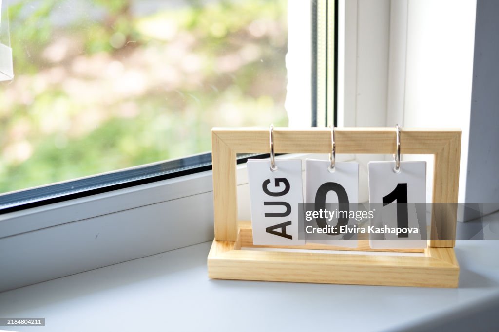 Wooden desk calendar with the date August 1 on the windowsill against the background of the window