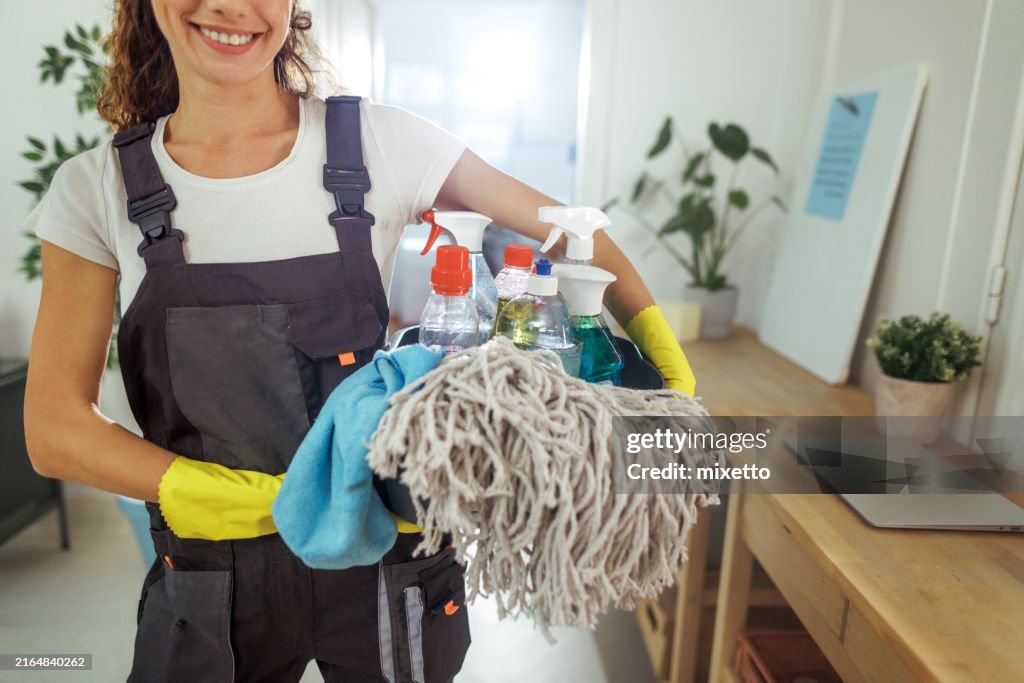 Cheerful young housewife holding bucket with cleaning supplies