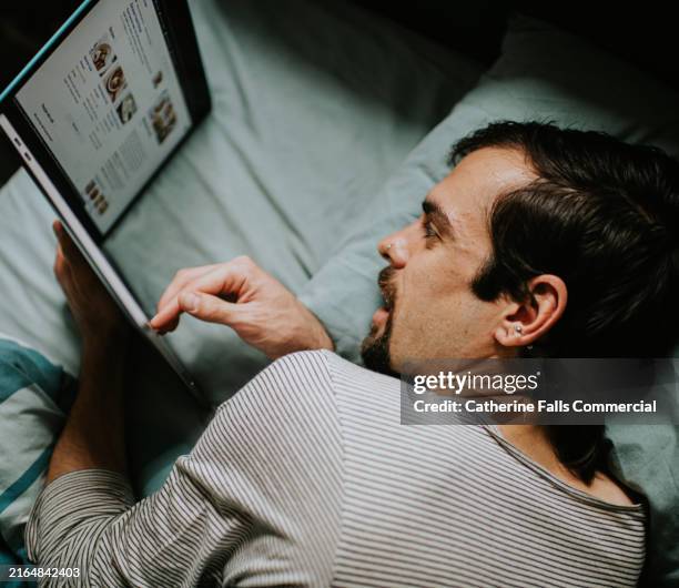 a man lies on his side in bed, propping his laptop sideways so he can look at it whilst lying down - laziness stock pictures, royalty-free photos & images