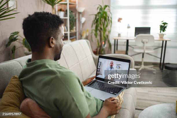 black man sitting on a sofa at home talking with his doctor on a video call over laptop - telemedicine stock pictures, royalty-free photos & images