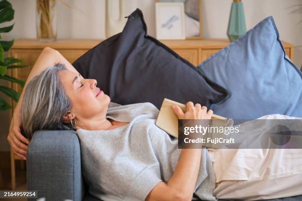 senior woman falling asleep while reading a book on the sofa - een dutje doen stockfoto's en -beelden