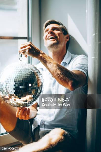 happy man joyfully holding a disco ball by a window, capturing a playful and bright moment. - eccentric stock pictures, royalty-free photos & images