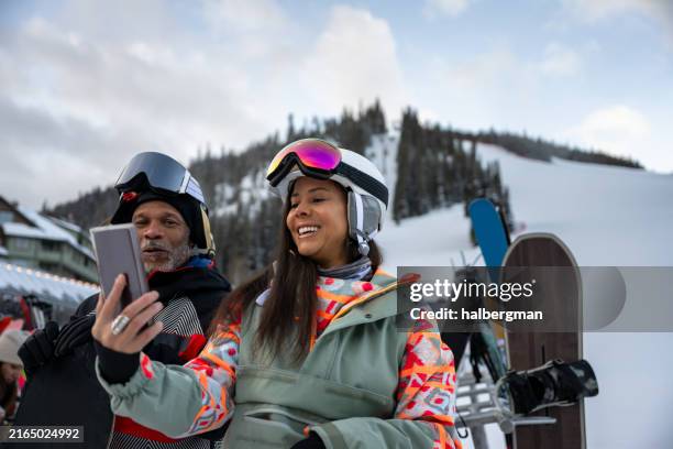 man and woman watching video on phone at base of ski slope - snow board stock pictures, royalty-free photos & images