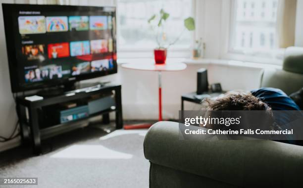 a man lies on a sofa, staring at a large, flatscreen tv displaying a choice of shows on a streaming platform - video on demand foto e immagini stock