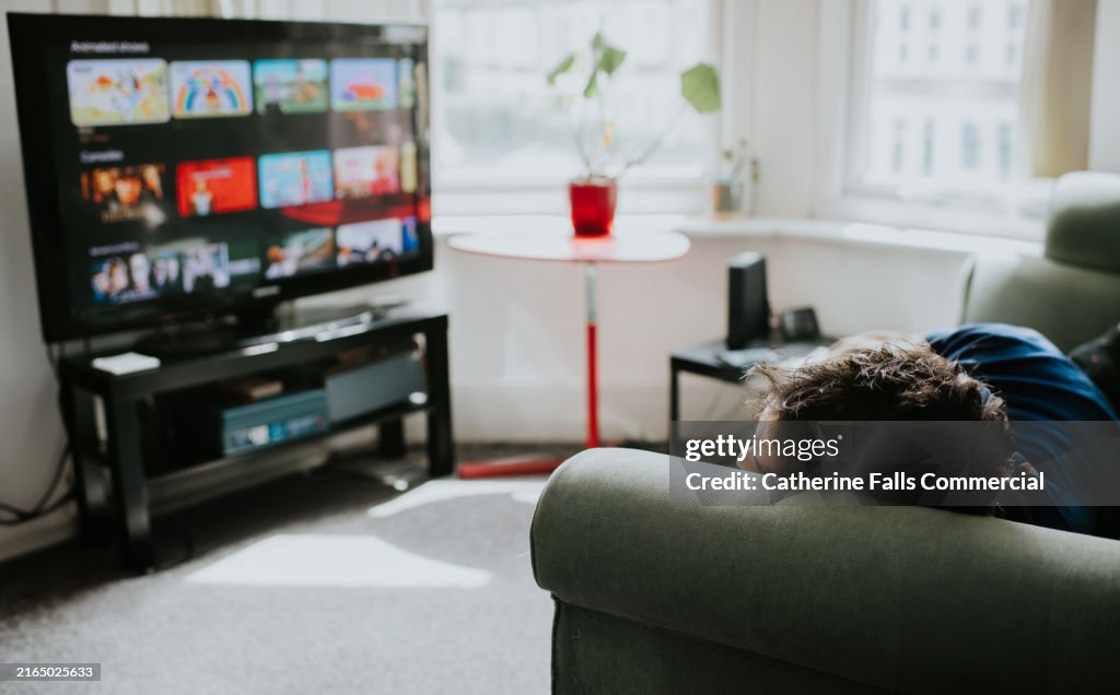 A man lies on a sofa, staring at a large, flatscreen TV displaying a choice of shows on a streaming platform