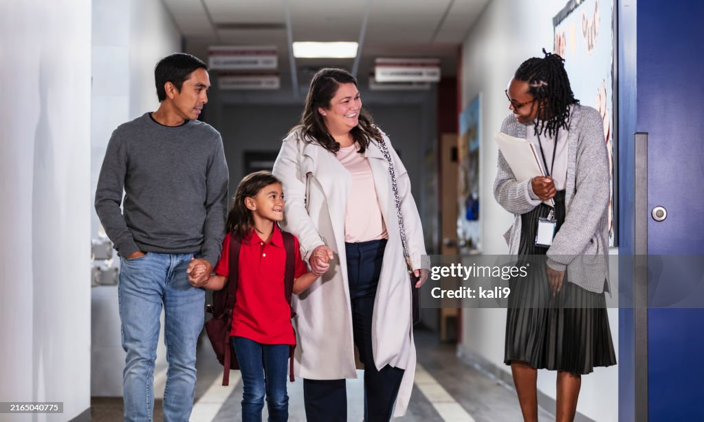 Parents bringing girl to school, with teacher in hallway