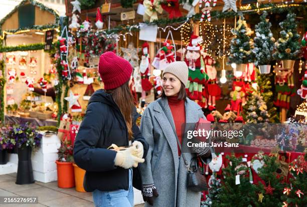 side view of smiling young female speaking friends in warm clothes looking at each other while carrying christmas gift packets in glove hands against market shops in daylight - julmarknad bildbanksfoton och bilder