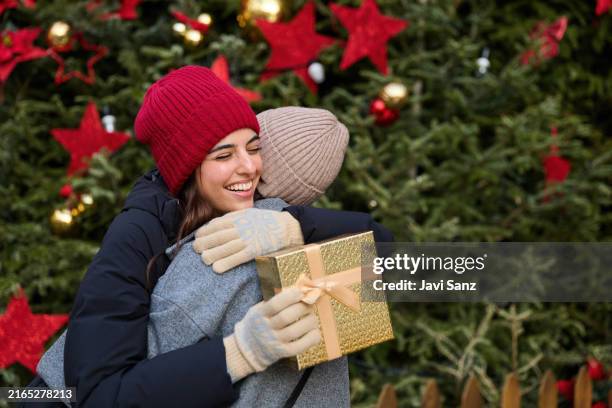 two happy friends exchanging christmas gift hugging near a christmas tree - exchanging gifts stock pictures, royalty-free photos & images