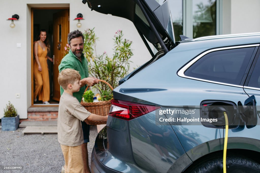 Father charging electric car after grocery shopping in supermarket. Plugging charger into electric vehicle in front of house.
