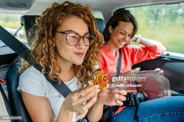 due amiche che usano un telefono e mangiano un bagel mentre guidano sul sedile posteriore dell'auto - stuzzicare foto e immagini stock