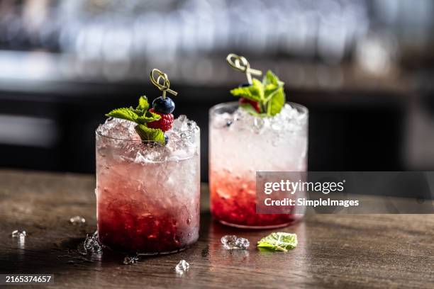 a traditional bramble drink made with gin, lemon juice, blackberry liqueur, berries and crushed ice on a bar counter in a night bar. - raspberry cocktail stock pictures, royalty-free photos & images