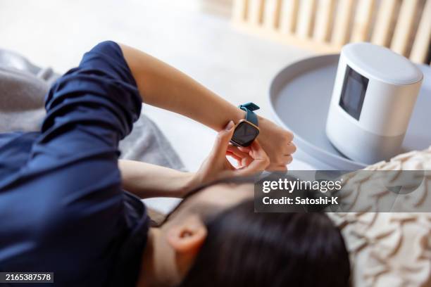 rear view of young woman lying down on bed, checking healthcare application on smart watch - smart speaker stock pictures, royalty-free photos & images