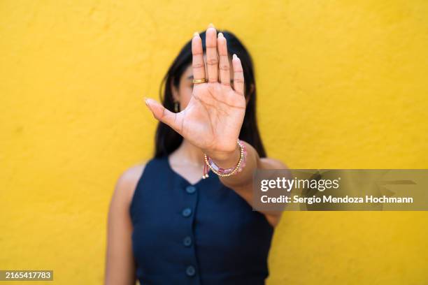 latino woman looking at the camera and gesturing to stop, hand obscuring her face - stopschild stock-fotos und bilder