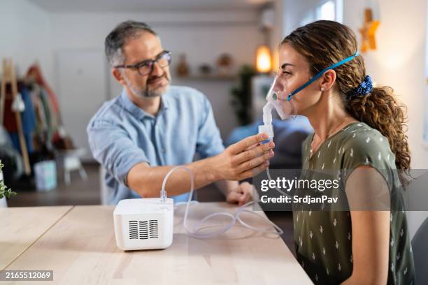 woman uses a nebulizer for respiratory therapy while her husband assists - nebulizer stock pictures, royalty-free photos & images