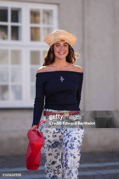Guest wears light brown hat, black off the shoulder long sleeve shirt, bright red belt, white pants with blue floral pattern/print, bright red...