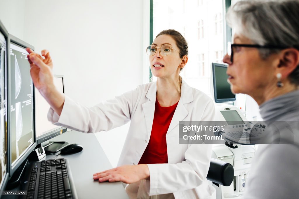 Young Female Doctor Pointing at Computer Screen While Explaining to Senior Patient