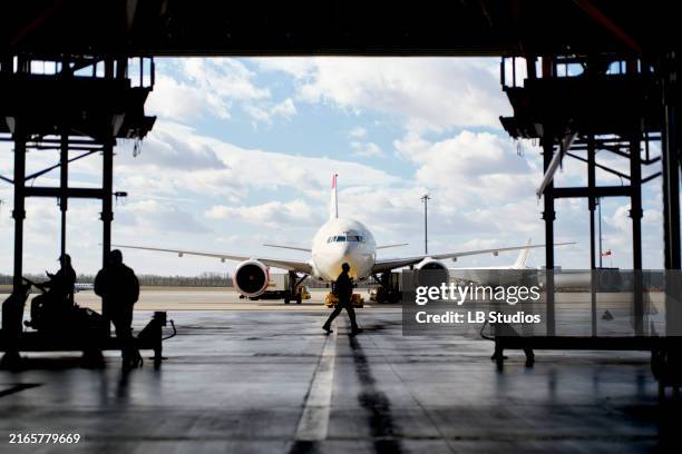 silhouetted figures work around an aircraft inside a hangar, with clear skies in the background - aerospace industry stock pictures, royalty-free photos & images