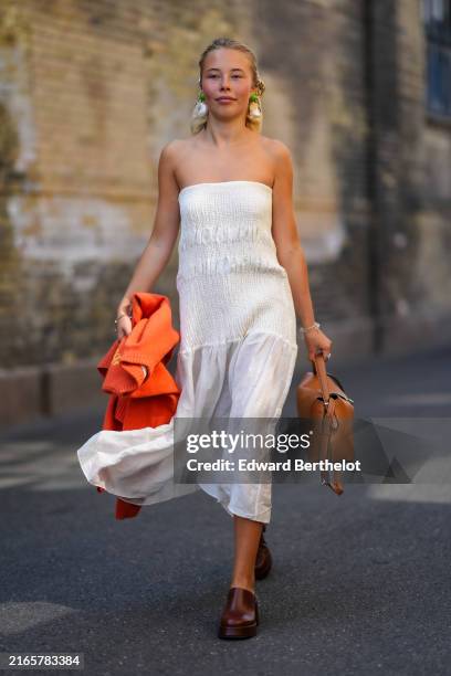 Guest wears wears beige black pattern head scarf, light green white earrings, white strapless ruffled midi dress, bright orange sweater in hand,...