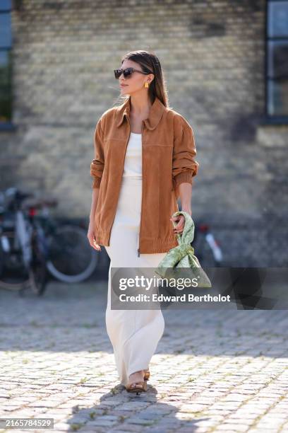 Guest wears dark brown sunglasses, gold earrings, white tank top, light brown oversized suede jacket, white maxi skirt with high slit, light green...