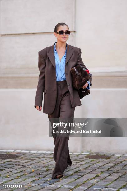 Darja Barannik wears dark brown sunglasses, silver earrings, light blue button up long sleeve shirt, dark brown oversized blazer jacket dark brown...