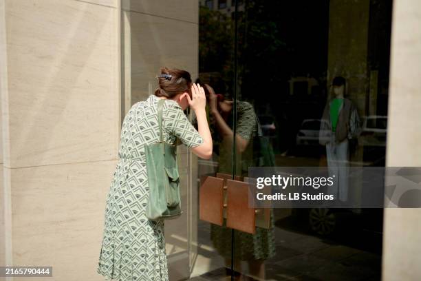 woman in a patterned dress looking at a storefront window, her reflection and a passerby visible in the glass - guardare le vetrine foto e immagini stock