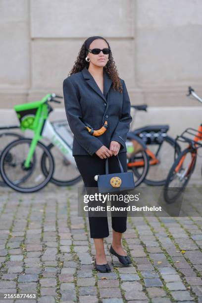 Guest wears black Prada sunglasses, black headband, silver earrings, black tailored blazer jacket, black leather banana holder, black Puppets and...