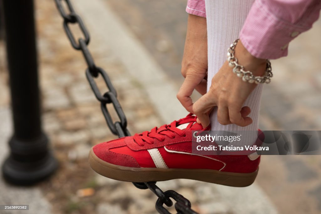 Leg of unseen woman tightening laces of vibrant red sneaker while resting foot on city street chain fence.