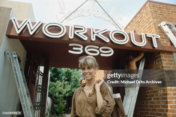 American actress Jane Fonda, wearing a brown checked blouse with the sleeves rolled up, outside her 'Workout' aerobics studio as a workman stands on...