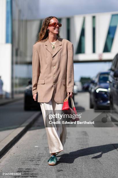 Guest wears red sunglasses, gold earrings, gold necklace, cream t-shirt, light brown oversized blazer jacket, cream loose pants, bright red leather...