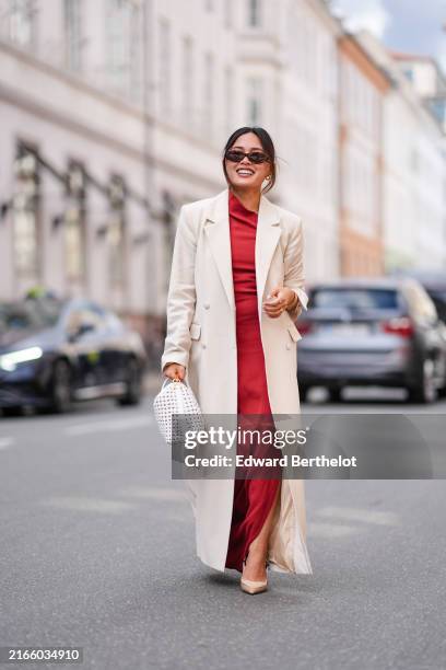 Guest wears dark brown sunglasses, gold earrings, cream blazer jacket coat, bright red asymmetrical maxi dress with high slit, gold bracelets, gold...