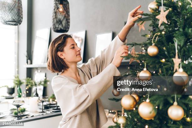 young woman is decorating christmas tree with many gold stars, balls and festive garland. new year celebration concept - decorar fotografías e imágenes de stock