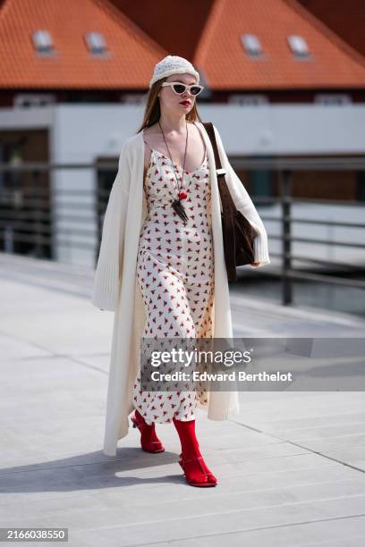 Guest wears white knit hat, cream sunglasses, black necklace red pendant, cream midi dress with red floral detail, cream cardigan trench sweater,...