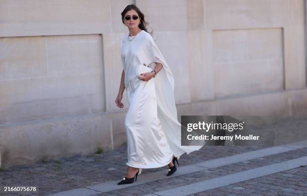 Darja Barannik is seen wearing a long white satin dress. She also wears black pumps with a white flower and white pearl, pearl earrings and pearl...