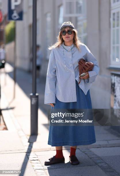 Fashion week guest is seen wearing a long blue denim skirt with an oversized blue and white striped blouse. She also wears large brown sunglasses, a...