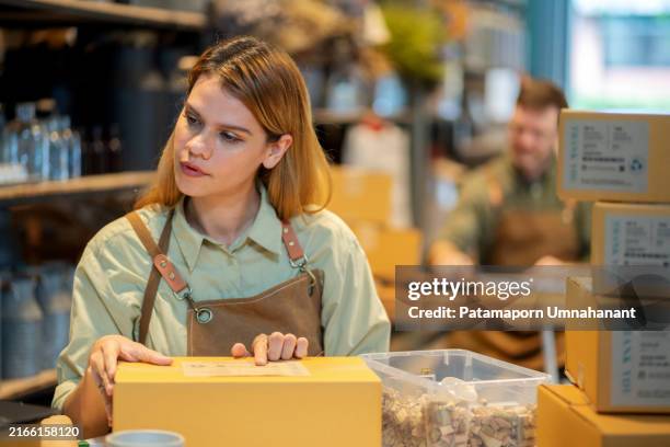 eine junge arbeiterin auf einer schürze verpackt die produkte sorgfältig in kartons mit natürlichen polstermaterialien in einem lagerhaus. ein kleines unternehmen, das sich auf nachhaltigkeit konzentriert, wobei der arbeiter sicherstellt, dass jede verp - biologisch abbaubar stock-fotos und bilder