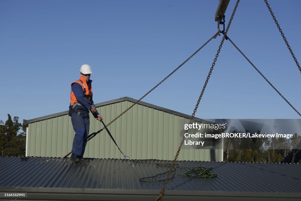 A builder wearing a safety harness helps to place the sling in place for a crane to lift a portable building