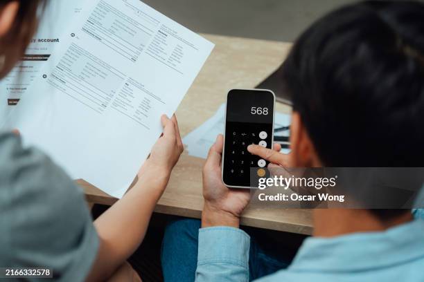 over the shoulder view of young asian couple going over financial bills and calculating budget with smartphone - expense stock pictures, royalty-free photos & images