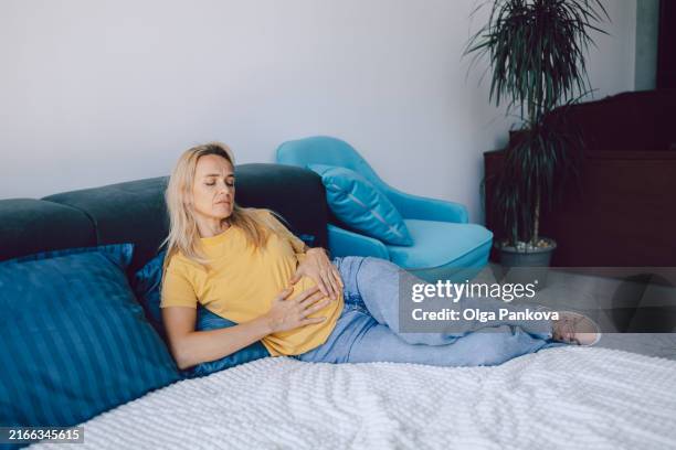 middle-aged woman holds her hands on her stomach while lying on a bed at home. pain, negative emotions and suffering. - irritable bowel syndrome stock pictures, royalty-free photos & images